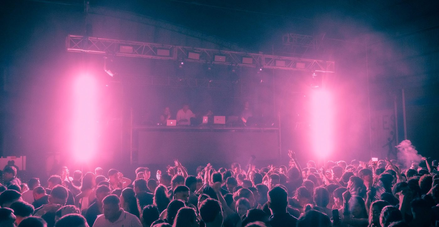 Público con las manos arriba en una discoteca, con luz rosa y la cabina del DJ al fondo
